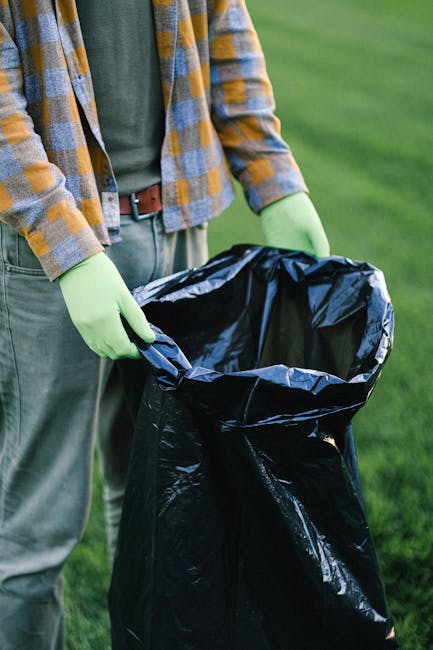 A person wearing a checkered shirt with shades of blue, yellow, and gray, along with green gloves, is holding a large, black high-visibility garbage bag open with both hands, standing outdoors on a gr
