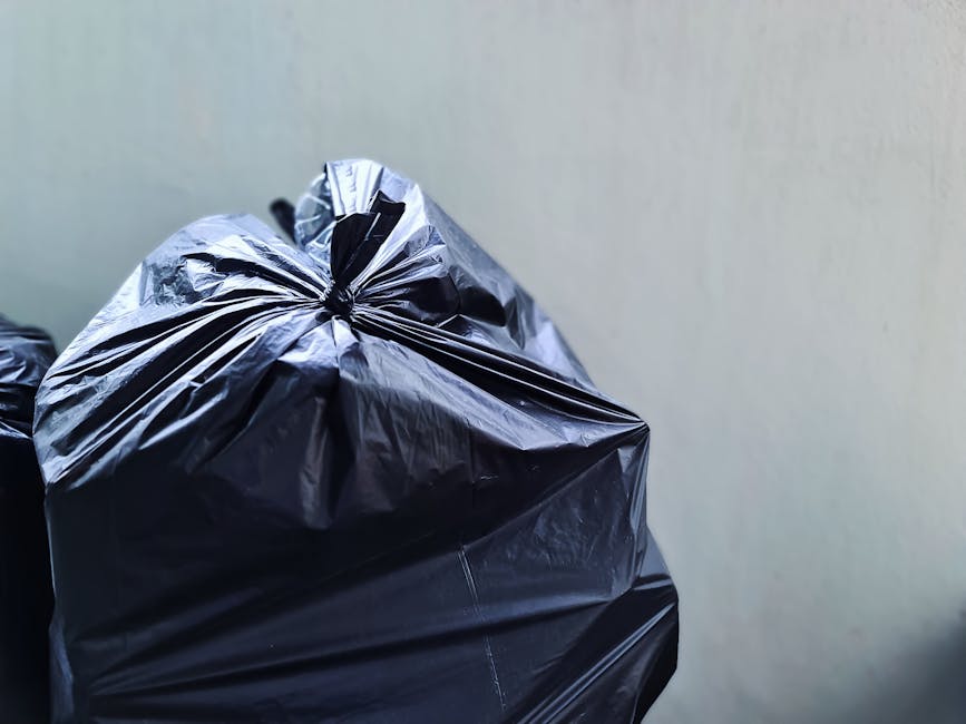 A person wearing a checkered shirt with shades of blue, yellow, and gray, along with green gloves, is holding a large, black high-visibility garbage bag open with both hands, standing outdoors on a gr